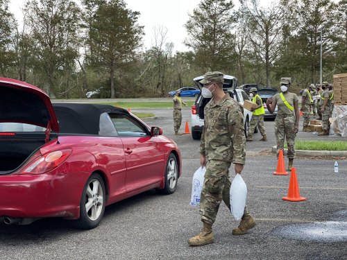 A soldier in a mask carries bags of supplies near vehicles in a parking lot, with others in the background.