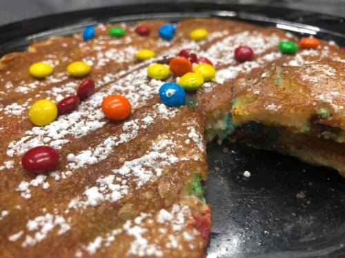 A close-up of a colorful pancake topped with powdered sugar and candy-coated chocolate pieces, with a slice cut out.
