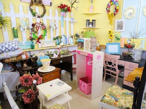 A cozy vintage shop filled with colorful decor, furniture, and floral arrangements on display against a bright yellow wall.