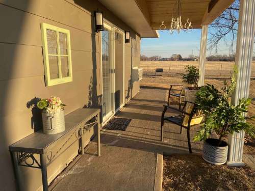 A cozy porch with a small table, chair, and potted plants, overlooking a grassy field under a clear sky.