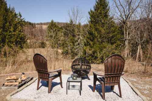 Two wooden chairs and a fire pit on a gravel patio, surrounded by trees and open fields under a clear blue sky.