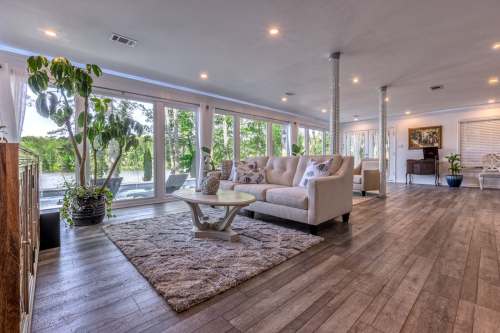 Bright, spacious living room with large windows, a cozy sofa, and wooden flooring, surrounded by greenery.