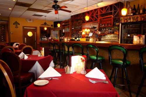 Cozy restaurant interior with red tablecloths, a bar area, and warm lighting, creating an inviting atmosphere.