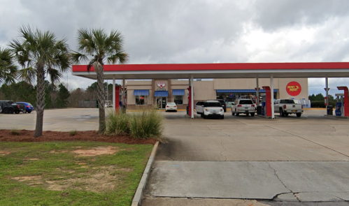 A gas station with a red roof, surrounded by palm trees and parked vehicles, under a cloudy sky.