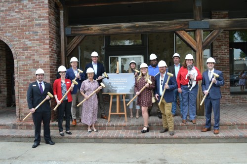 A group of people in hard hats poses with mallets in front of a building, celebrating a groundbreaking event.