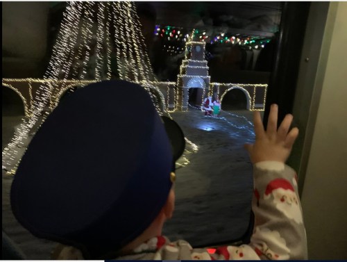 A child in festive pajamas waves at a colorful holiday light display outside a window.