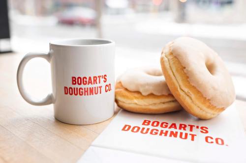 A white mug and two glazed donuts with "Bogart's Doughnut Co." printed on them, placed on a wooden surface.
