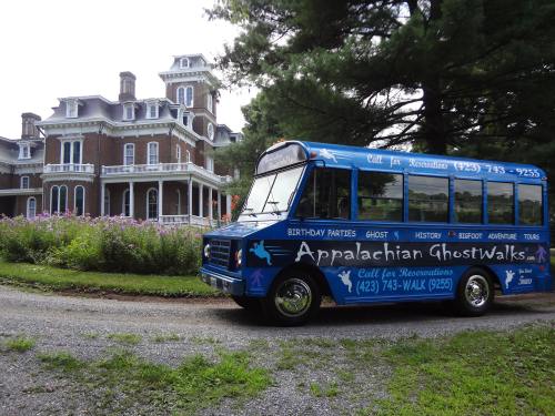 A blue tour bus parked in front of a large historic mansion, surrounded by trees and flowers.