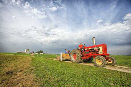 A red tractor pulls a cart along a grassy path, with a cloudy sky and farm buildings in the background.