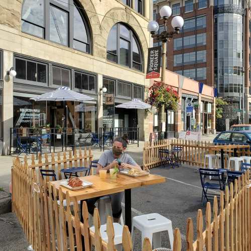 Outdoor dining area with wooden barriers, tables, and a person enjoying a meal in a city setting.