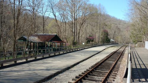 A quiet train station with a wooden platform and tracks, surrounded by trees and mountains under a clear blue sky.