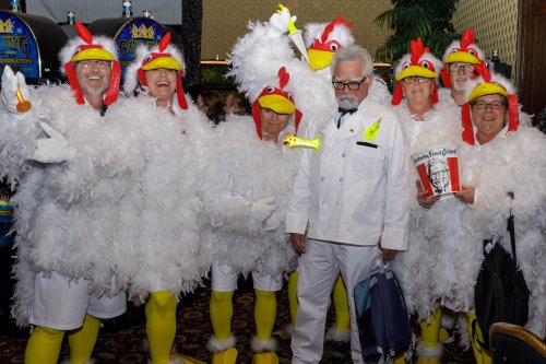 A group of people in chicken costumes poses with a man in a white suit, resembling a famous chicken brand mascot.