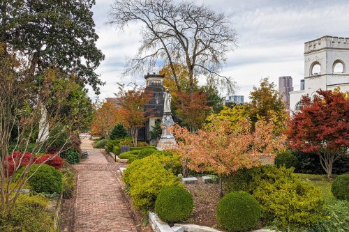 A serene garden path lined with colorful autumn foliage and manicured shrubs, leading to a statue and buildings in the background.