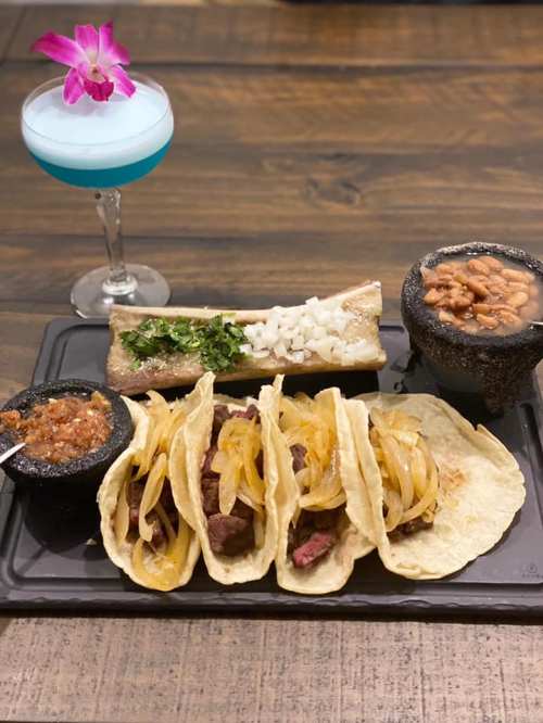A plate of tacos with grilled meat and onions, served with beans, salsa, and a blue cocktail garnished with a flower.