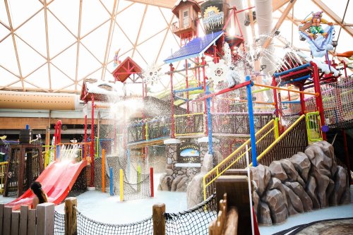 A colorful indoor water park with slides, play structures, and water features under a geometric glass roof.