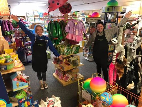 Two smiling women in aprons stand in a colorful toy store, surrounded by shelves of toys and playful items.