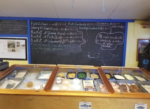 A seafood menu board with prices, featuring items like fish and chips, shrimp, and clam chowder, above a display case.