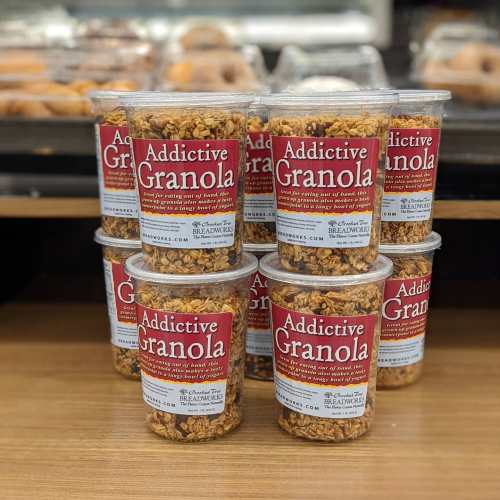 A stack of clear cups filled with granola, labeled "Addictive Granola," displayed on a wooden surface.