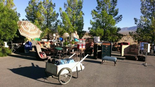 A yard sale with various items like furniture, tools, and decor set up outdoors under trees and a large umbrella.