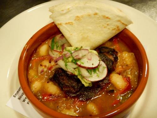 A bowl of hearty stew with meat, vegetables, and a side of flatbread, garnished with radishes and herbs.