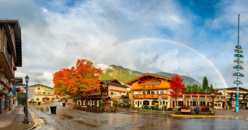 A picturesque town square with colorful autumn trees, shops, and a rainbow arching over the mountains in the background.