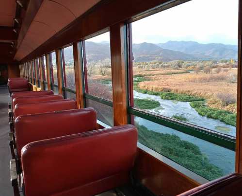 Interior of a train car with red seats, showcasing a scenic view of a river and mountains through large windows.