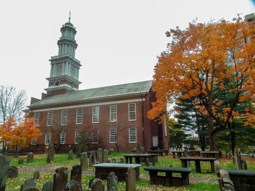A historic brick church with a tall clock tower, surrounded by autumn trees and a graveyard with stone markers.