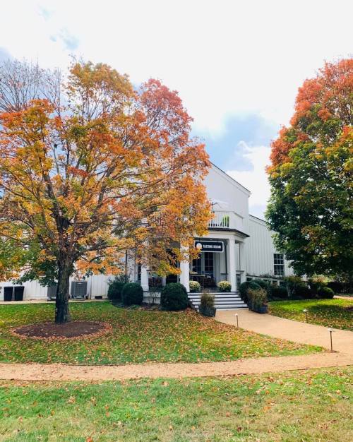 A white building surrounded by colorful autumn trees and a grassy path, under a partly cloudy sky.