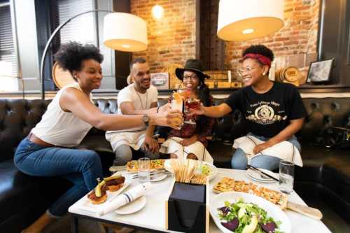 Four friends toast with drinks at a table filled with food in a cozy restaurant setting.