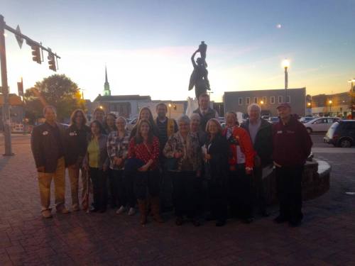 A group of people poses together near a statue at dusk, with buildings and a church steeple in the background.