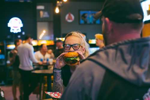 A woman with glasses smiles while holding a large burger in a lively restaurant setting.