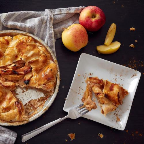 A freshly baked apple pie on a striped cloth, with a slice on a plate and whole apples nearby.