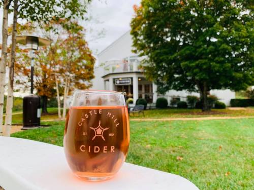 A glass of cider sits on a ledge, with a scenic view of a building and colorful autumn trees in the background.