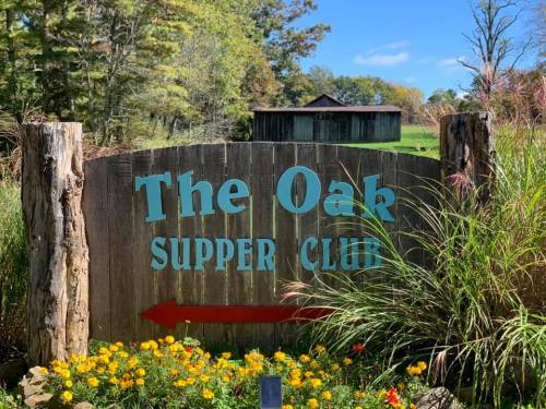 Sign for "The Oak Supper Club" with a directional arrow, surrounded by colorful flowers and greenery.