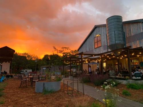 A rustic building with outdoor seating, surrounded by greenery, under a vibrant sunset sky.