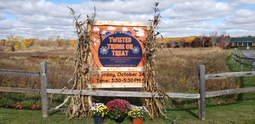 Sign for "Twisted Trunk or Treat" event with date, time, and autumn scenery in the background.