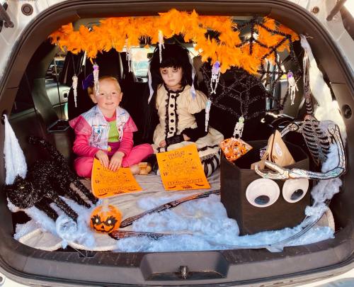 Two children in a decorated car trunk for Halloween, surrounded by spider webs, orange decorations, and candy.