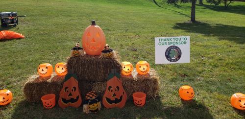 A festive display of pumpkins and jack-o'-lanterns on hay bales, with a sign thanking the sponsor in the background.