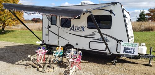 A parked RV with an awning, surrounded by colorful chairs and a sign thanking the Banes Family for sponsorship.