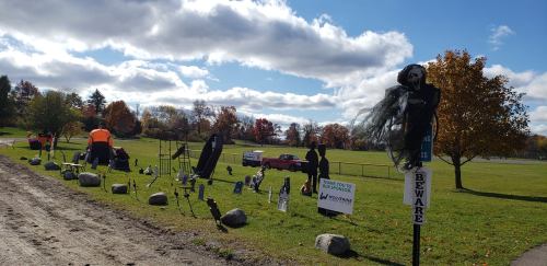A park scene with Halloween decorations, including a skeleton and various spooky props, under a cloudy sky.