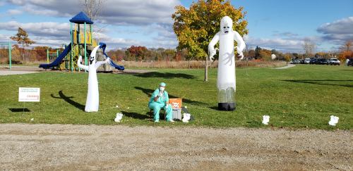 A park scene with inflatable ghosts, a costumed figure in scrubs, and a playground in the background under a blue sky.