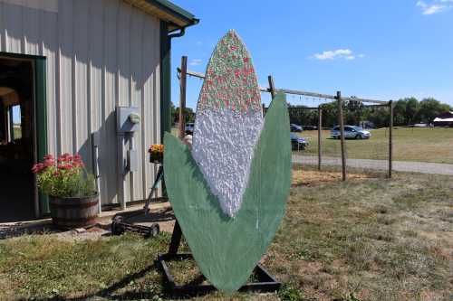 A large, painted corn cob sculpture stands outside a building, with a blue sky and grassy area in the background.