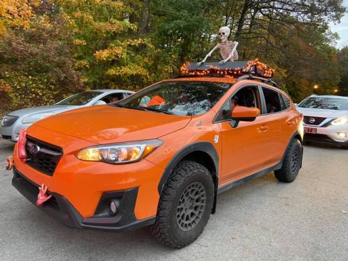 An orange SUV decorated for Halloween with a skeleton on the roof and festive lights. Trees with autumn leaves in the background.