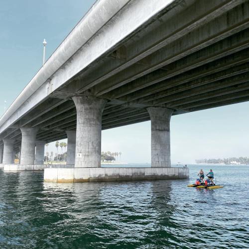 A concrete bridge spans over calm water, with kayakers paddling beneath it on a sunny day.