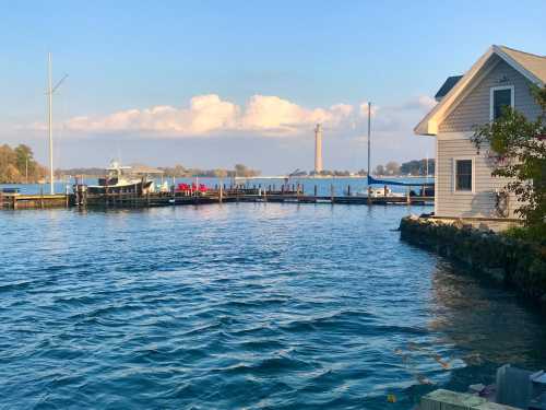 A serene waterfront scene with a dock, boats, and a lighthouse in the distance under a clear blue sky.