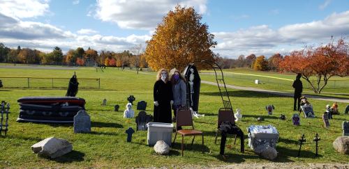 Two people stand in a graveyard setting with tombstones and Halloween decorations, surrounded by autumn trees.