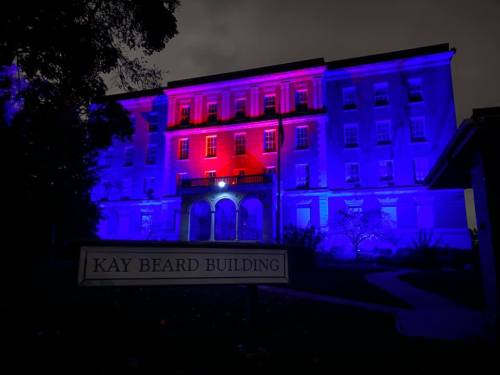 The Kay Beard Building illuminated in red and blue lights at night, surrounded by dark trees.