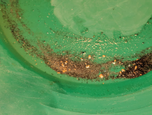 Close-up of a green pan with black sand and small gold flakes scattered along the bottom.