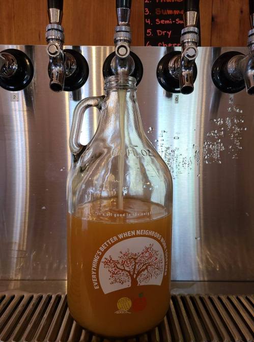 A glass growler filled with a light amber beverage sits in front of beer taps on a metal wall.