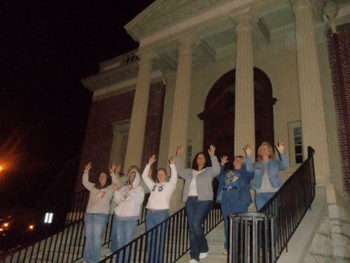 A group of six women stands on the steps of a building at night, raising their hands in celebration.